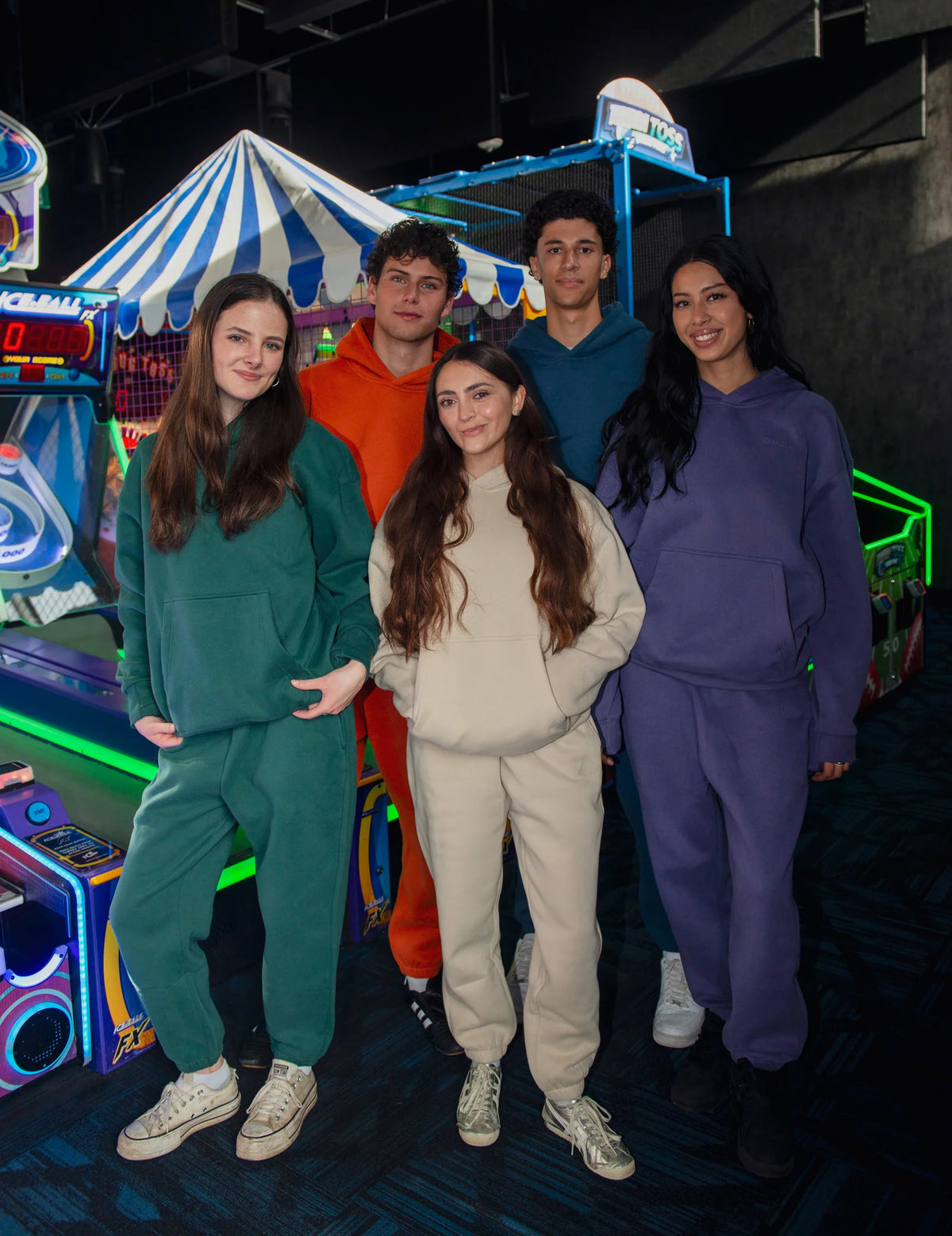 Five adults wearing Soothe hoodies and matching sweatpants in green, orange, blue, cream and purple at an arcade.